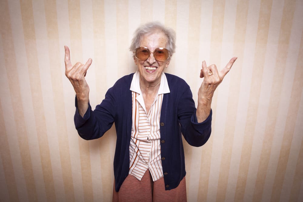 Ältere Dame mit Sonnenbrille, die lächelnd die Hände mit dem „Rock on" Zeichen in die Höhe hält.
